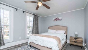 Une chambre à coucher avec un lit, un ventilateur de plafond, une table de chevet et une fenêtre avec des rideaux.