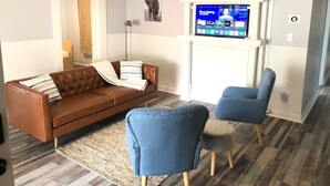 Un salon avec un canapé en cuir marron, deux fauteuils bleus, une cheminée blanche et un téléviseur fixé au-dessus.