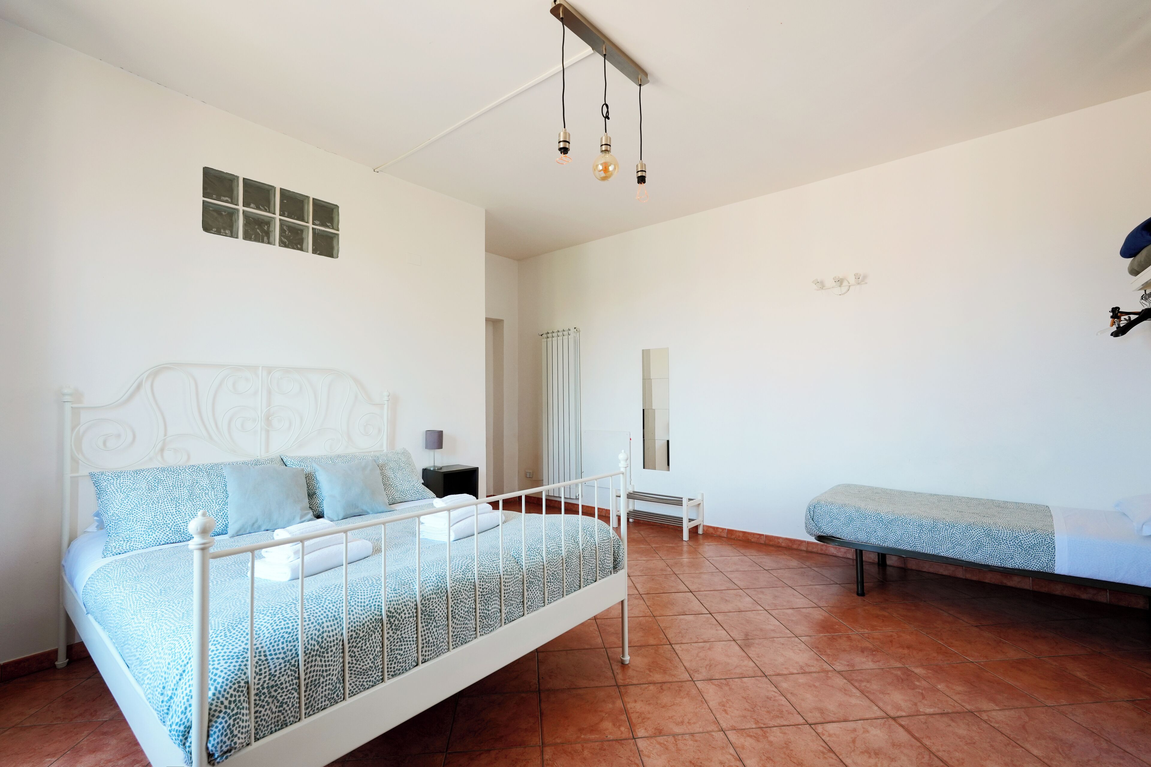 Una camera da letto con un letto in ferro bianco, una coperta a motivi, un comodino e una panca.
