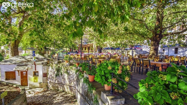 An outdoor seating area with tables and chairs under trees, a stone wall, and a small building with a door and window. - Milies Creekside Studio (South Pelion)