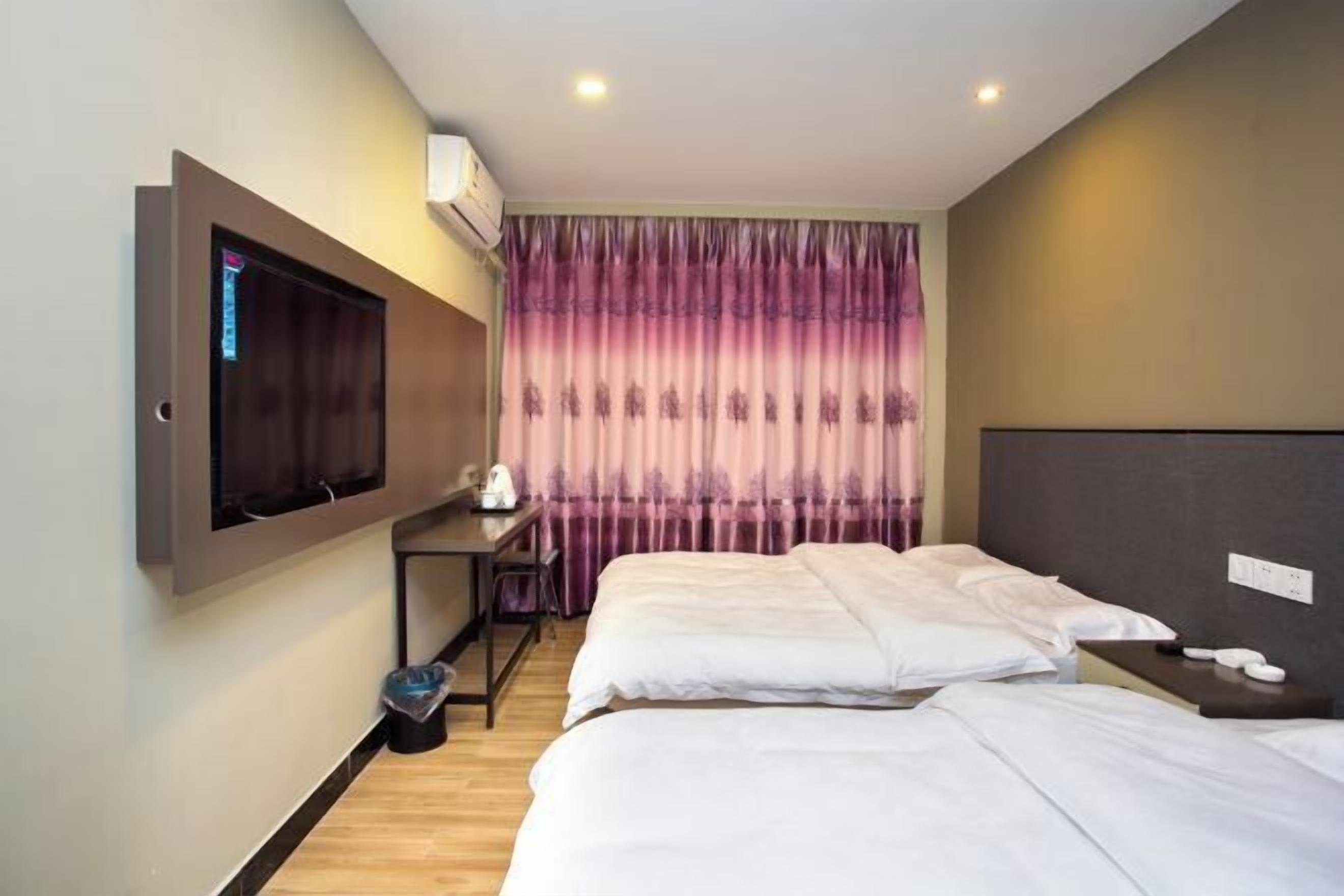 A hotel room with two beds, a desk with a chair, a TV mounted on the wall, and a curtain with a purple pattern.