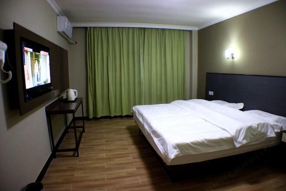 A hotel room with a bed, a TV on the wall, a small table with a cup, and a curtain.