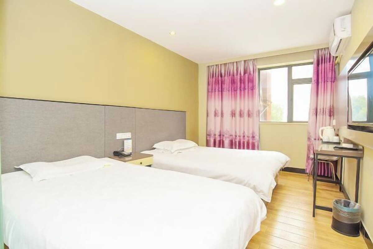 A hotel room with two single beds, a desk with a chair, a window with curtains, and an air conditioning unit.