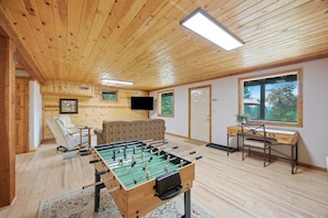 A cozy room with a foosball table, a sofa, a television, and a window with a view of trees.
