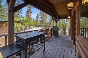 An outdoor grill area with a wooden deck, a metal grill, and a view of a forest.
