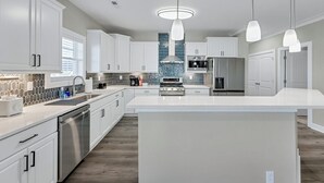 Una cocina moderna con armarios blancos, una isla central, electrodomésticos de acero inoxidable e iluminación colgante.