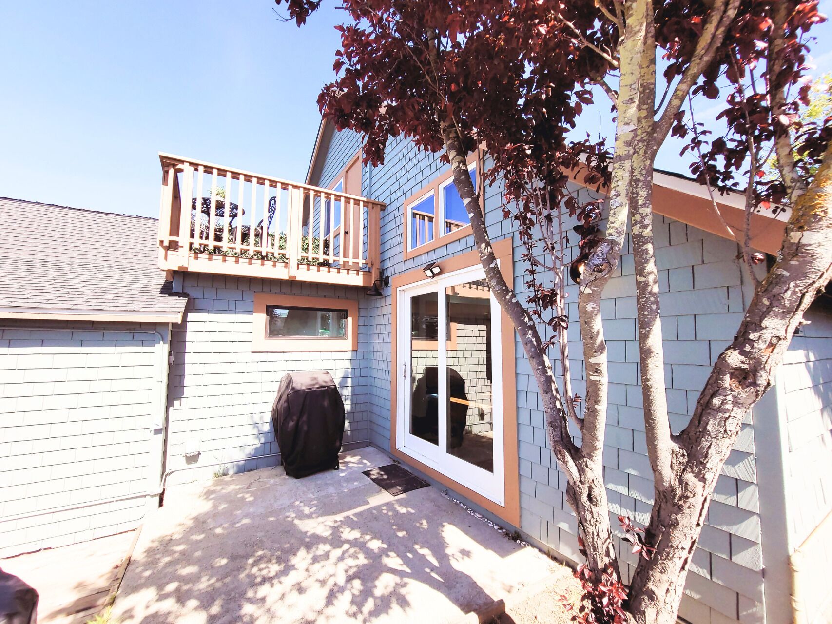 A residential backyard with a covered grill, a wooden deck, and a tree.