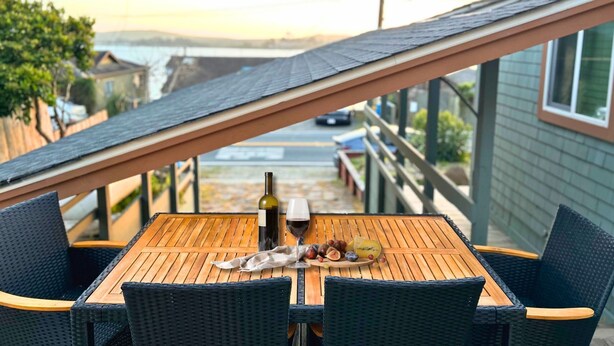 Una mesa de madera con sillas de mimbre, una botella de vino y una bandeja de frutas en un balcón con vistas a una calle y una masa de agua.