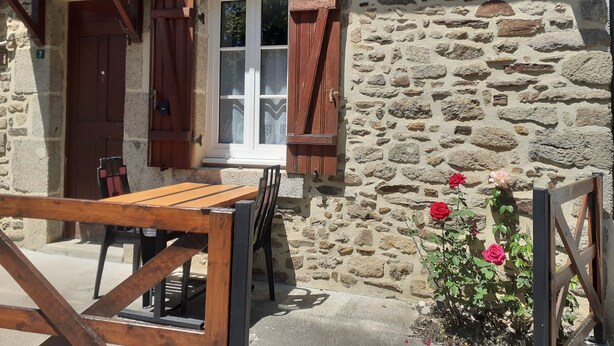 A stone building with a wooden table and chairs outside, a window with curtains, and a rose bush. - QUIETLY SITUATED VACATION HOME (Azerables)