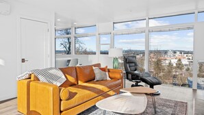 Een moderne woonkamer met een groot raam, een leren bank, een zwarte fauteuil en een kleine ronde tafel.