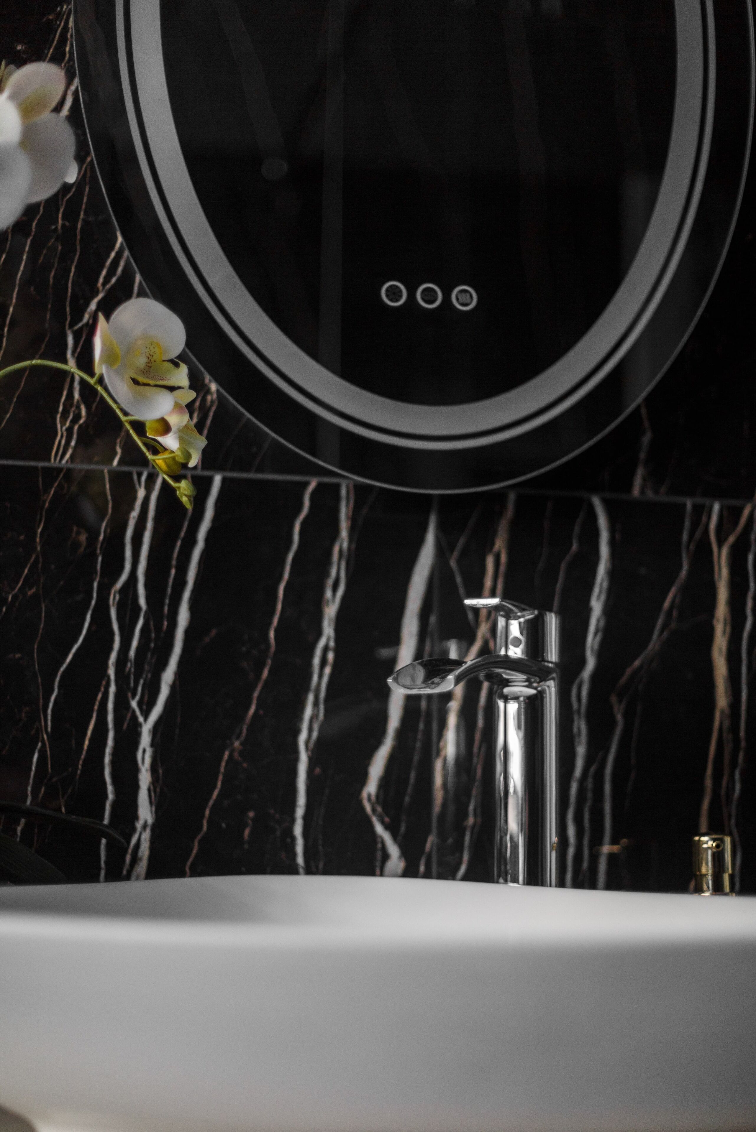 A modern bathroom with a marble backsplash, a round mirror, and a chrome faucet.