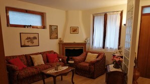 Una sala de estar con sofá, sillón y una mesita. Hay una chimenea, una ventana con cortinas y una puerta.