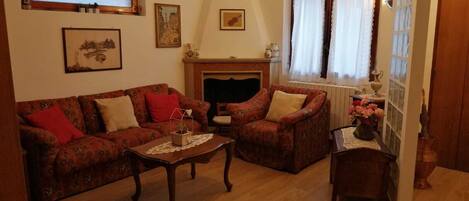 A living room with a sofa, armchair, and a small table. There is a fireplace, a window with curtains, and a door.