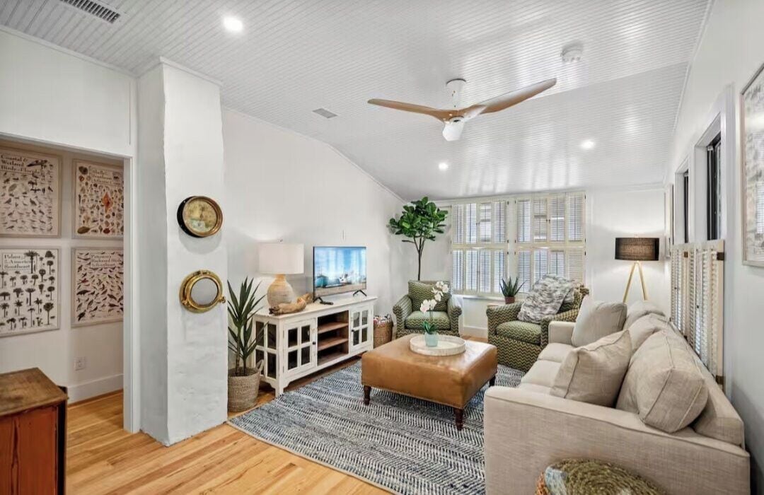 A spacious living room with a high ceiling, a ceiling fan, a television, and a large window providing natural light.