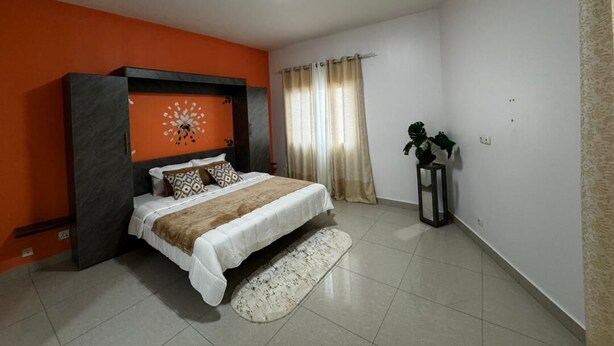 A bedroom with a bed, an air conditioner, a window with curtains, and a small plant. - Sofana Residence – 2 Bedroom Apartment (Dakar)