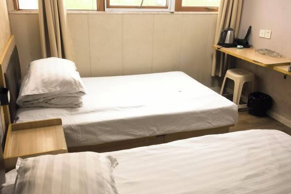 A small, single-room accommodation with two single beds, a wooden nightstand, a small stool, a shelf with a kettle, and a radiator.