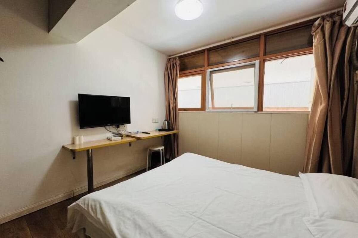 A hotel room with a bed, a desk with a flat-screen TV, a window with curtains, and a radiator.