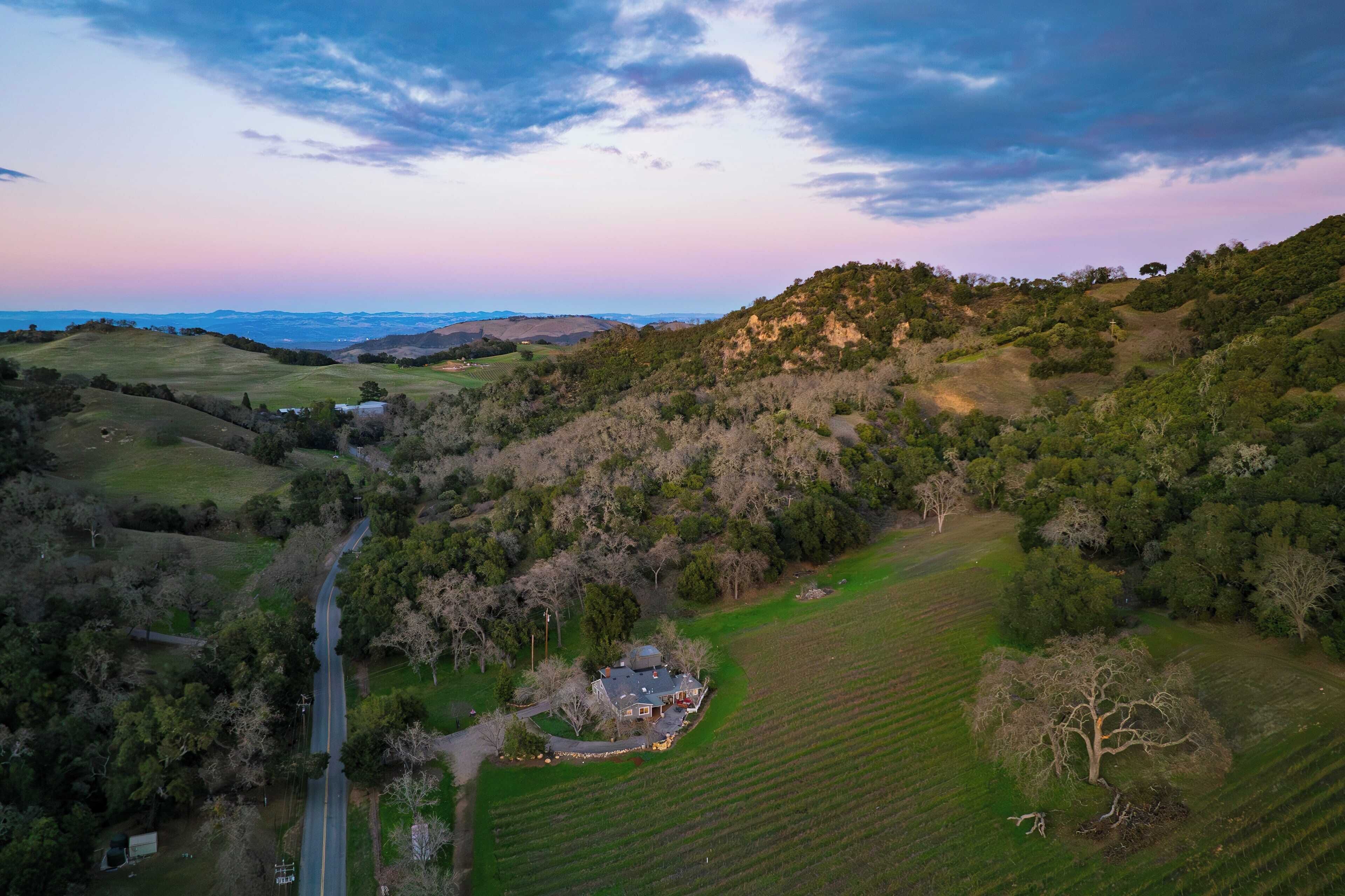 An aerial view of a rural landscape with rolling hills, a winding road, and a house surrounded by fields and trees.