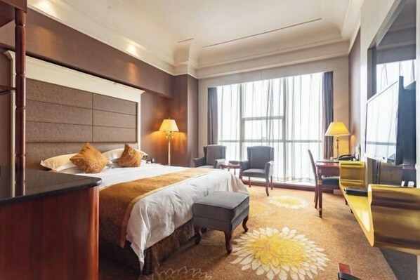 Ein Hotelzimmer mit einem großen Bett, zwei Nachttischlampen, einem Schreibtisch mit Stuhl und einem großen Fenster mit Vorhängen.
