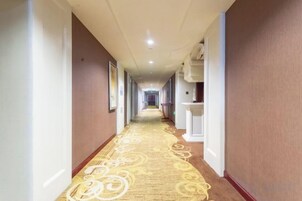 A hotel corridor with a patterned carpet, framed pictures on the walls, and a long hallway leading to a door at the end.