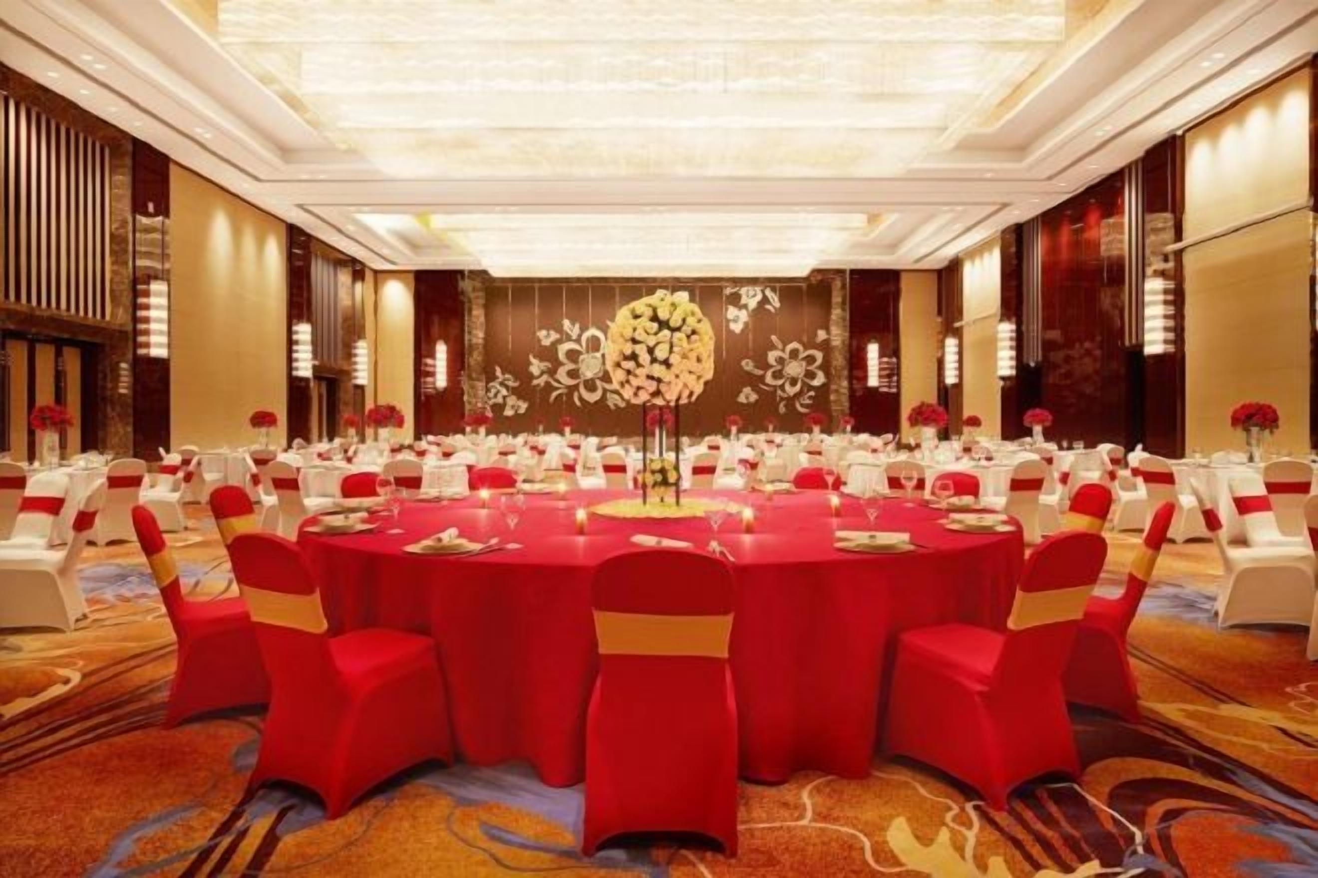 Un salón de banquetes con mesas redondas, sillas rojas y una decoración floral en la pared.