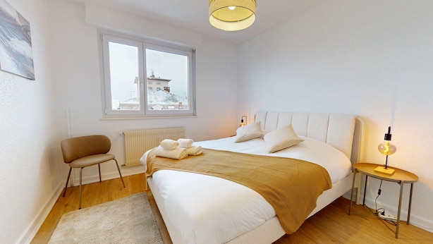 A modern bedroom with a large bed, a chair, a bedside table with a lamp, and a view of buildings through a window. - La belle étape + 1 parking (Colmar)