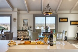 Ein helles Zimmer mit einem großen Fenster, einem Esstisch mit Snacks und Wein und Blick auf die Landschaft.
