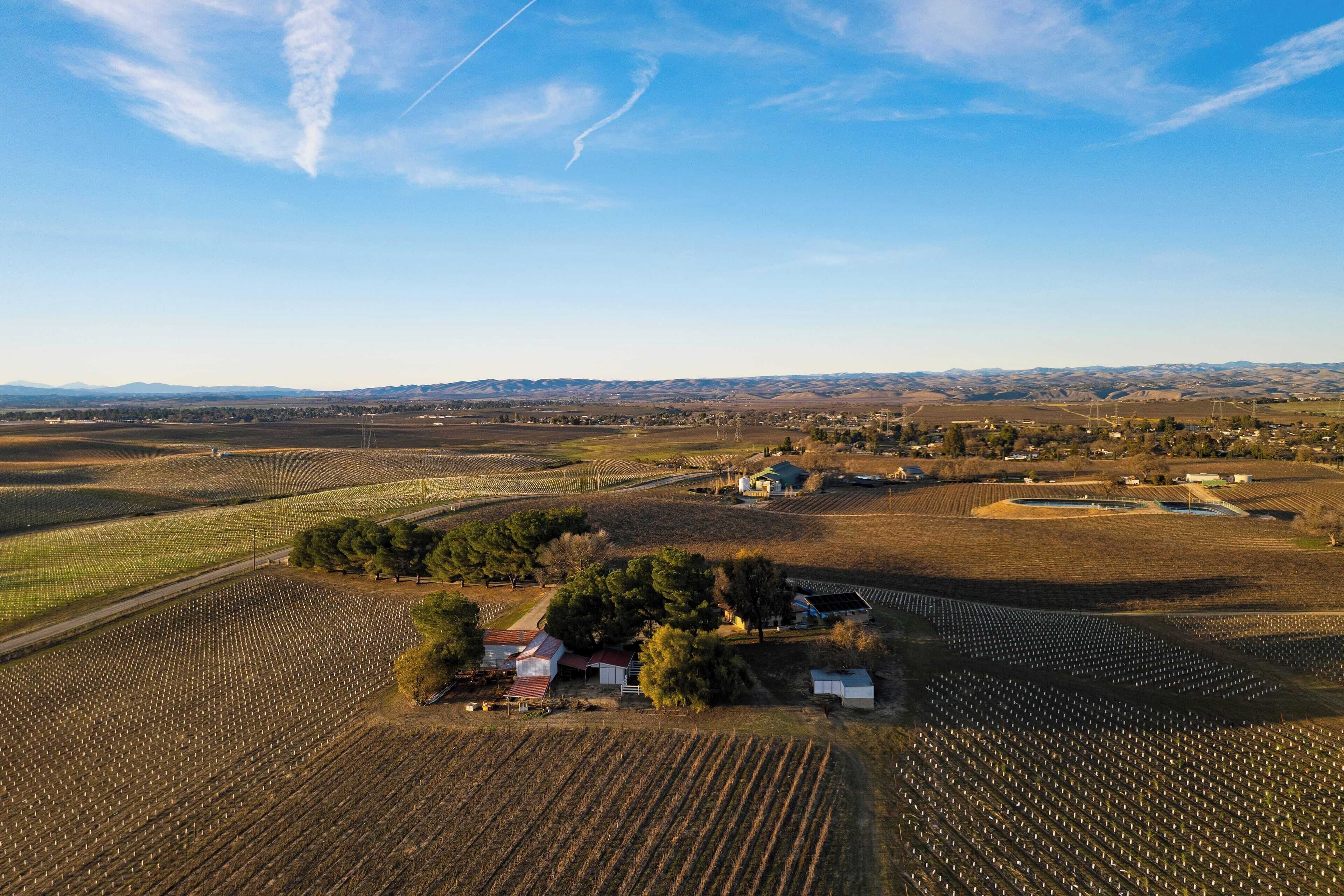 Vista aérea de un paisaje rural con campos, un pequeño grupo de edificios y montañas a lo lejos.