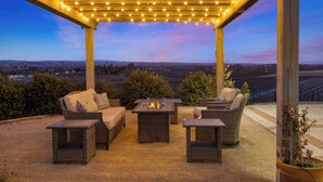Un patio exterior con mobiliario de mimbre, un hogar para fuego y una pérgola con luces colgantes.