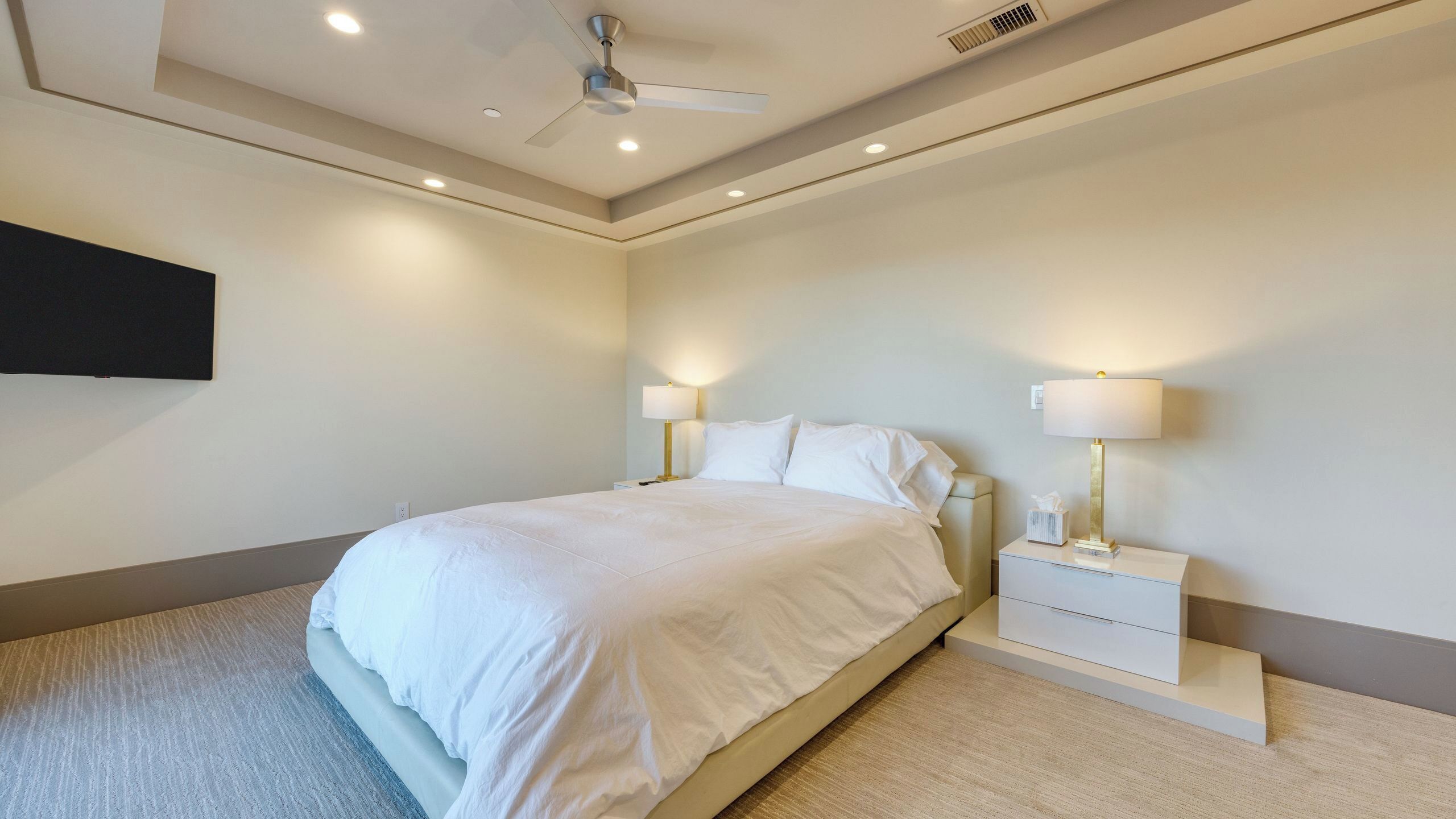 Un dormitorio con una cama, dos mesitas de noche con lámparas, un ventilador de techo y un televisor montado.