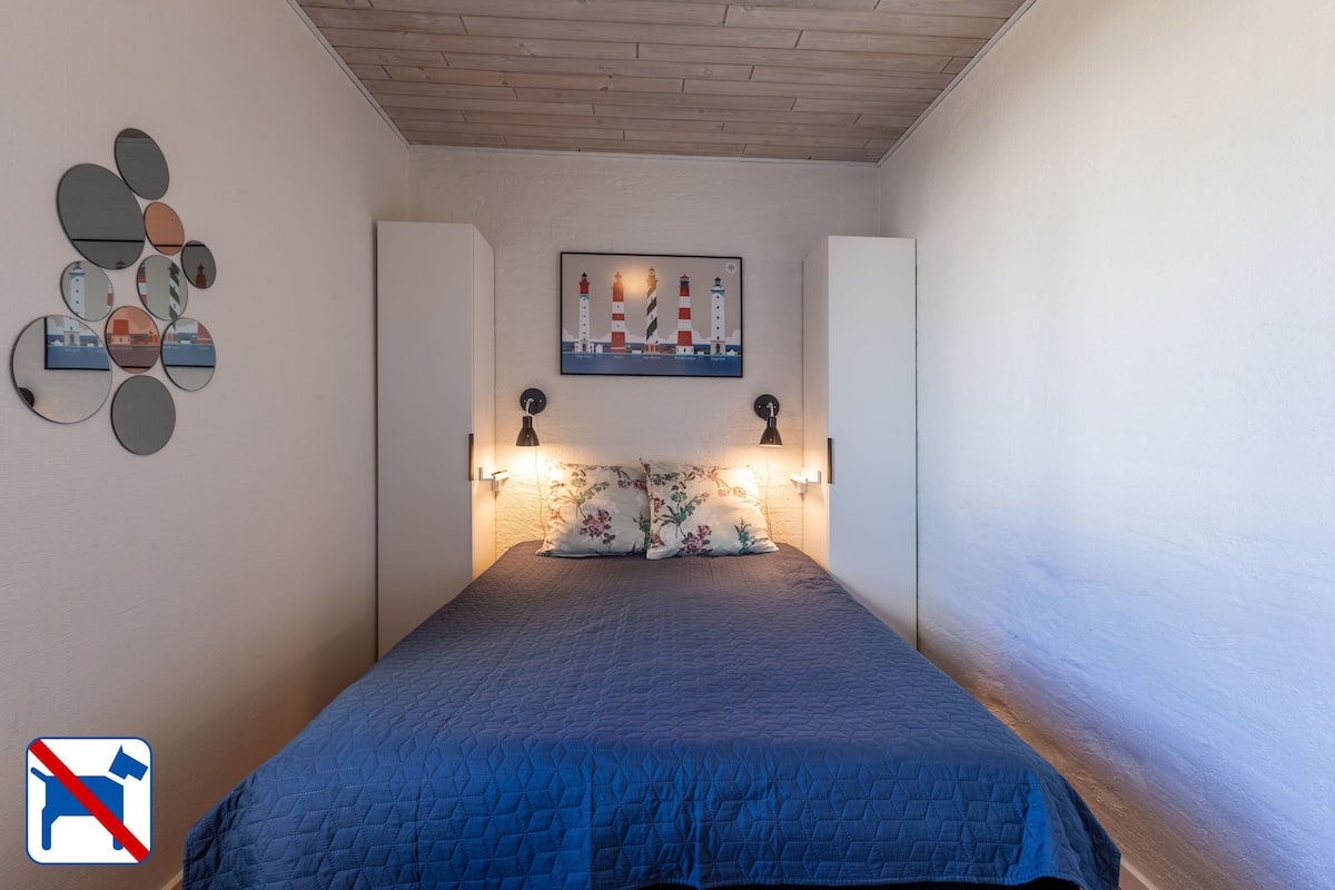 Ein Schlafzimmer mit blauer, gesteppter Bettdecke, einem an der Wand befestigten Spiegel, einem gerahmten Bild von Leuchttürmen und zwei Wandlampen.