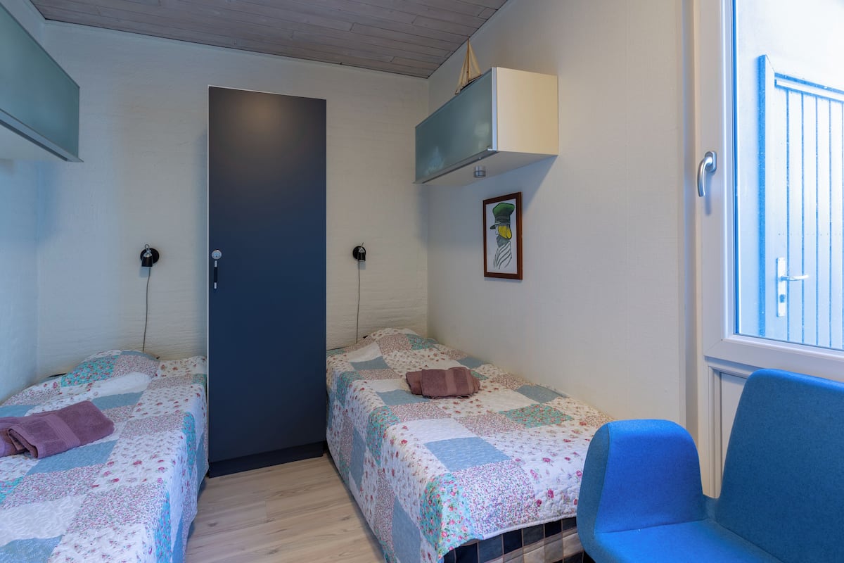 Ein Zimmer mit zwei Einzelbetten, einem blauen Stuhl, einer Tür und einem kleinen Fenster.