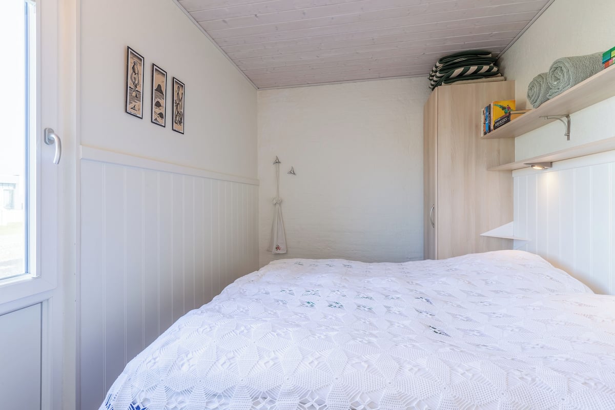 Ein Schlafzimmer mit weißer Bettdecke, einem Holzschrank und einem Regal mit Büchern und aufgerollten Matten.