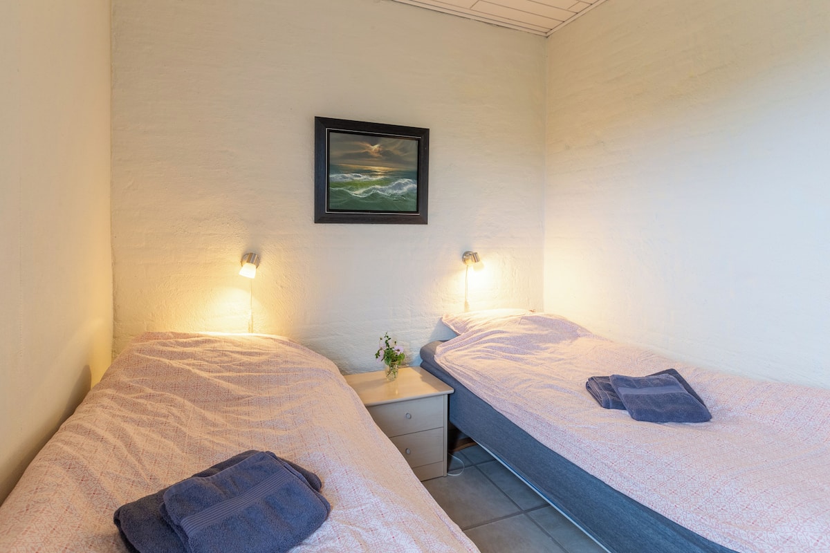 Ein Doppelzimmer mit zwei Betten, jeweils mit Kissen und zusammengelegtem Handtuch. Ein kleiner Nachttisch mit Lampe und ein gerahmtes Bild an der Wand.