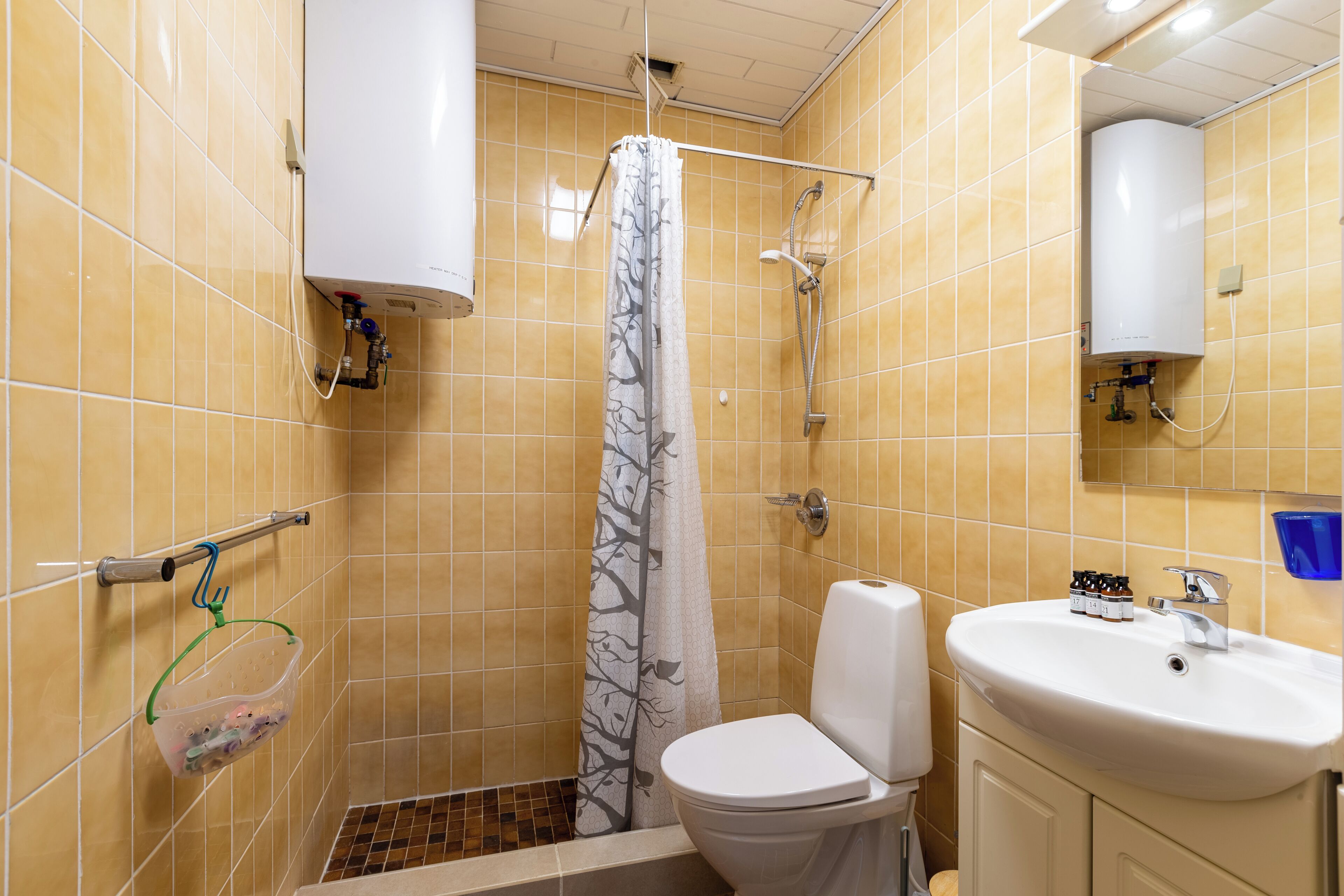A bathroom with yellow tiles, a toilet, a sink, a shower with a tree-patterned shower curtain, and a towel rack.