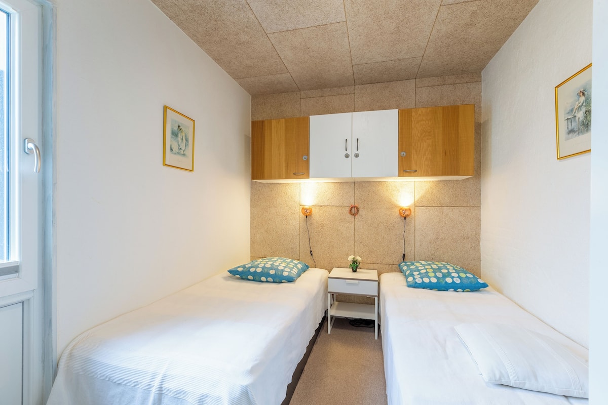Ein kleines, sauberes Zimmer mit zwei Einzelbetten, einem kleinen Tisch und einem Schrank über den Betten.