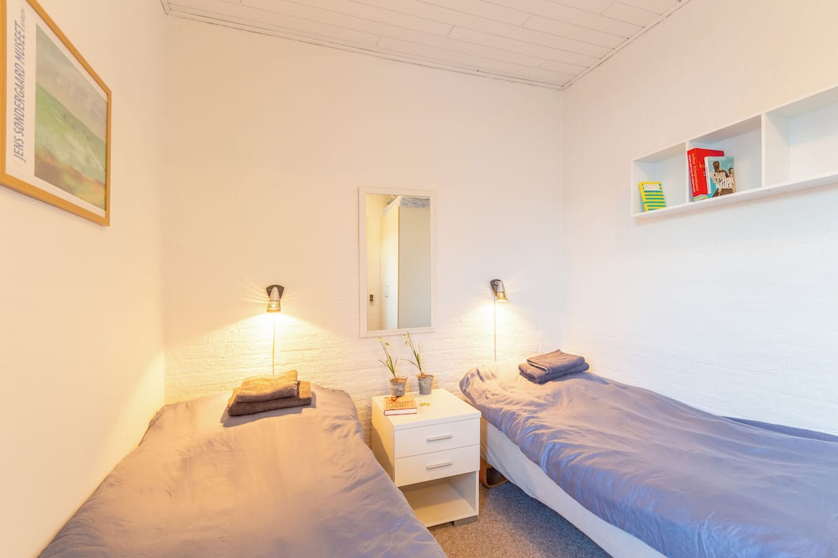 Ein Schlafzimmer mit zwei Einzelbetten, einem Nachttisch mit Lampe, einem Spiegel und einem Regal mit Büchern.