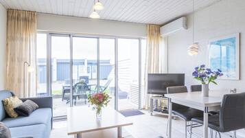 Una sala moderna con un sofá, una mesa de comedor y un televisor. Hay un ventanal y una puerta que da a un espacio exterior.