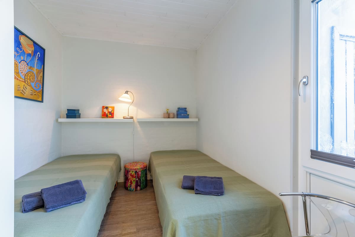 Ein kleines, sauberes Zimmer mit zwei Einzelbetten, einem kleinen Tisch mit einer Lampe und einem Regal über dem Bett.