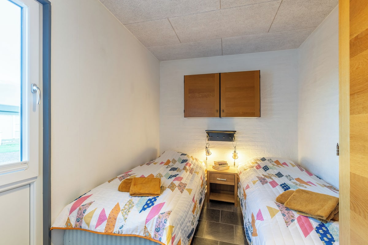 Ein kleines, gut beleuchtetes Schlafzimmer mit zwei Einzelbetten, einem Holzschrank, einem Nachttisch mit Lampe und einem Fenster für natürliches Licht.