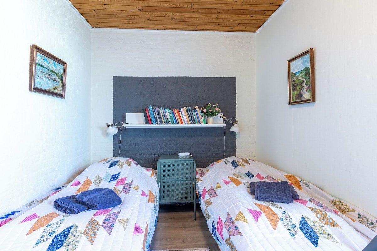 Ein Schlafzimmer mit zwei Einzelbetten, einem Nachttisch mit Büchern und einer Lampe sowie einer Wandablage mit dekorativen Gegenständen.
