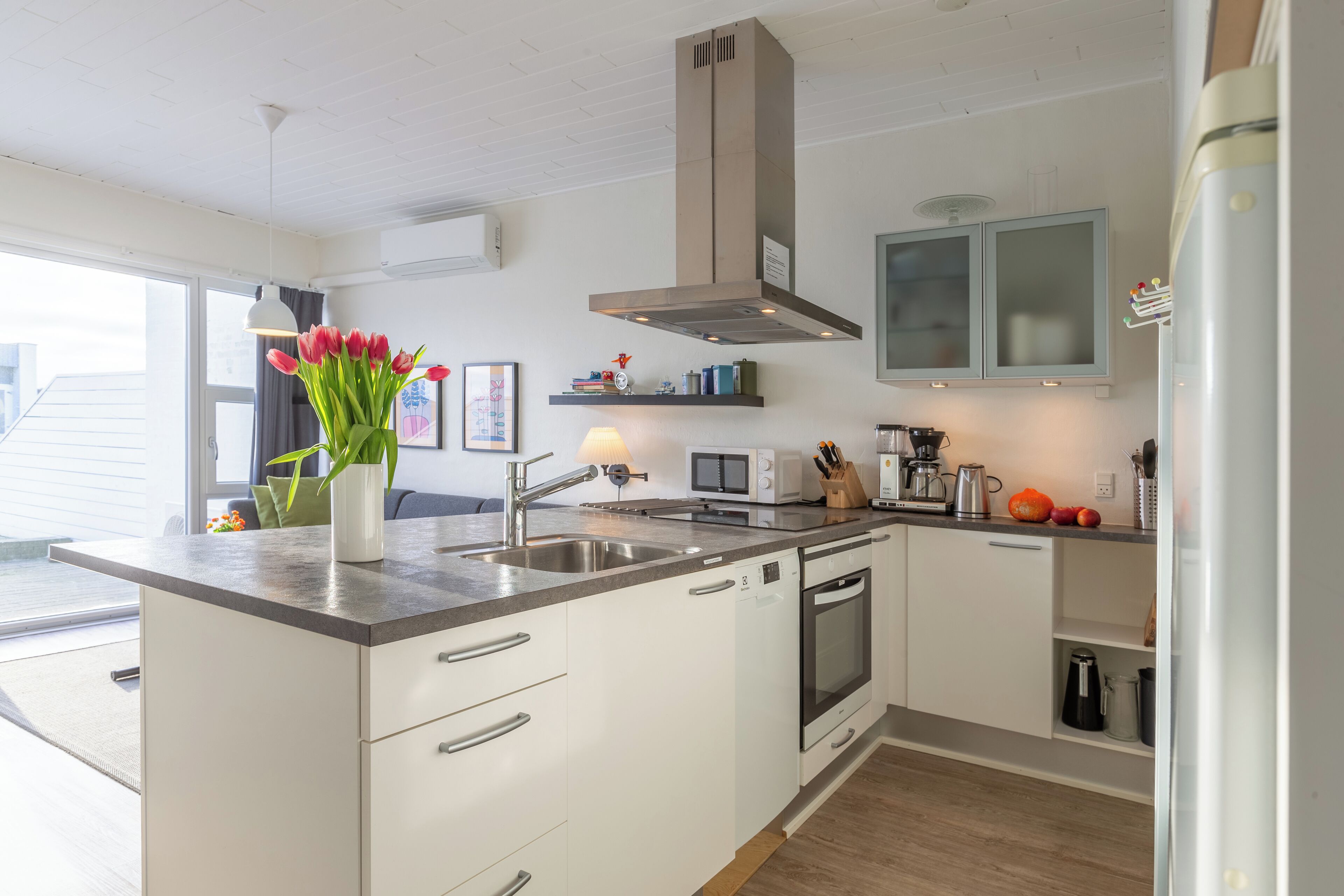A modern kitchen with white cabinets, a stainless steel sink, and a built-in oven. There is a vase with red flowers on the counter, and a microwave above the sink.