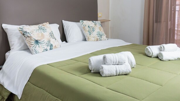 Un lit bien fait, avec une couvre-lit verte, des oreillers blancs et un coussin décoratif à motifs floraux. On peut voir des serviettes sur le lit et un tableau accroché au mur.