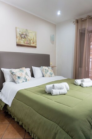 Een netjes opgemaakt bed met een groene plaid, witte kussens en een sierkussen met bloemenprint. Handdoeken liggen op het bed en aan de muur hangt een schilderij.