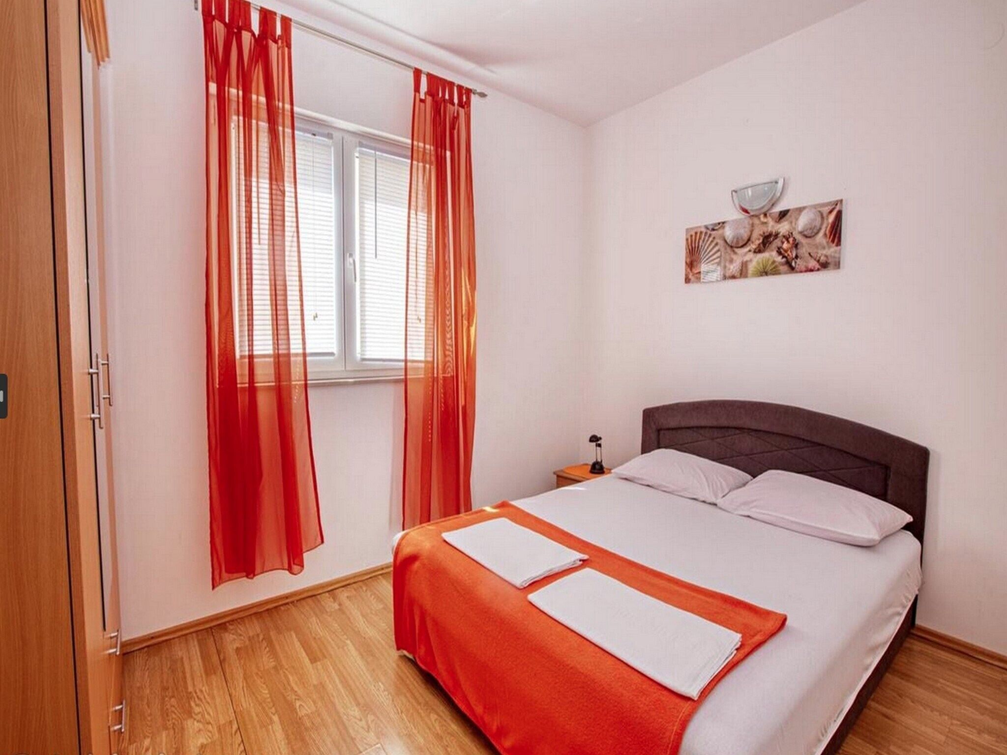 Une chambre à coucher avec un lit, une fenêtre ornée de rideaux rouges, une table de chevet avec une lampe et un tableau accroché au mur.