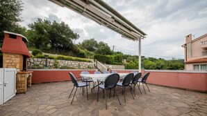 Eine Terrasse im Freien mit Steinboden, einem Tisch mit Stühlen, einem Grill und einer Pergola.