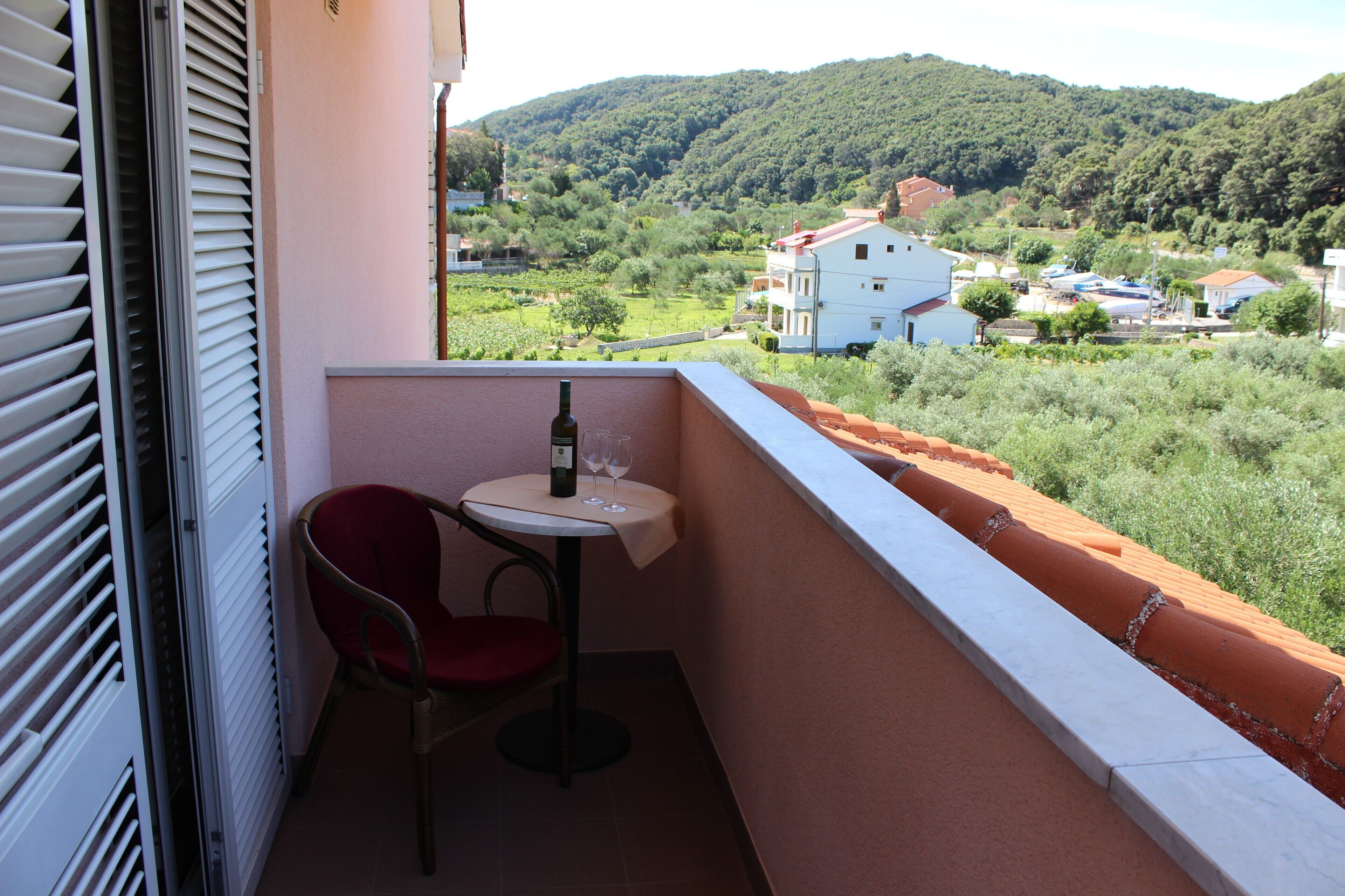 A balcony with a table and chair, overlooking a green landscape with buildings in the distance.