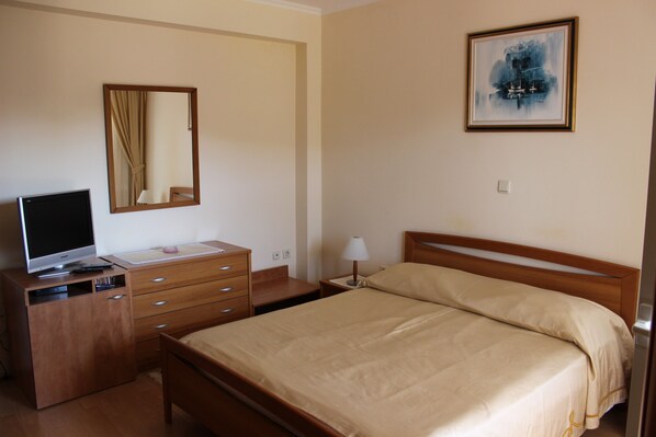 A hotel room with a bed, a TV on a wooden dresser, a mirror, a painting on the wall, and a nightstand with a lamp.