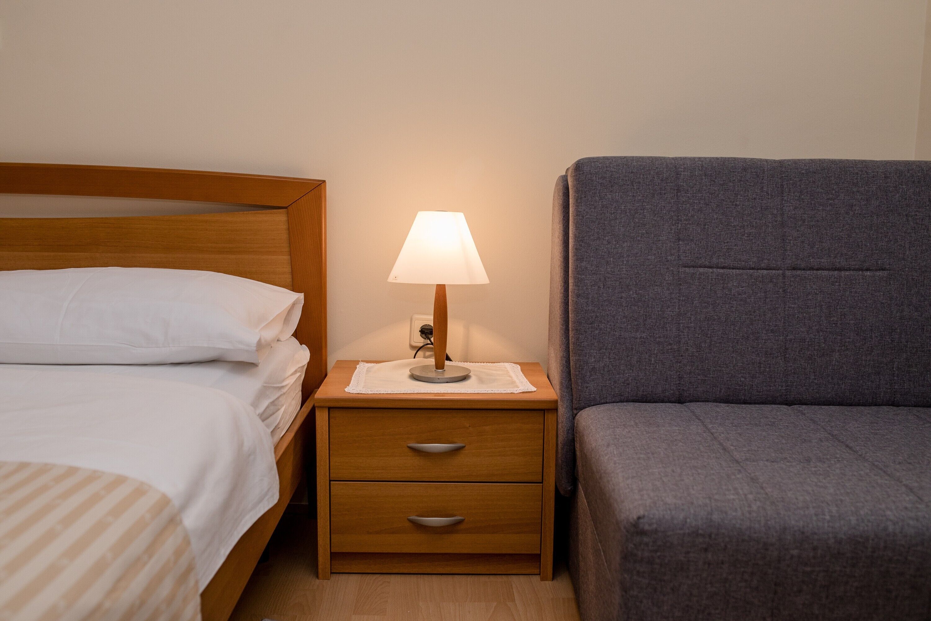 Een netjes opgemaakt bed met witte lakens, een houten nachtkastje met een lamp, en een grijze gestoffeerde stoel.