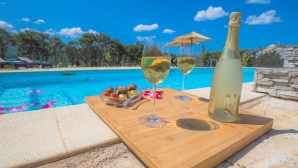 A wooden table with a bottle of white wine, two glasses, and a bowl of snacks by a pool.